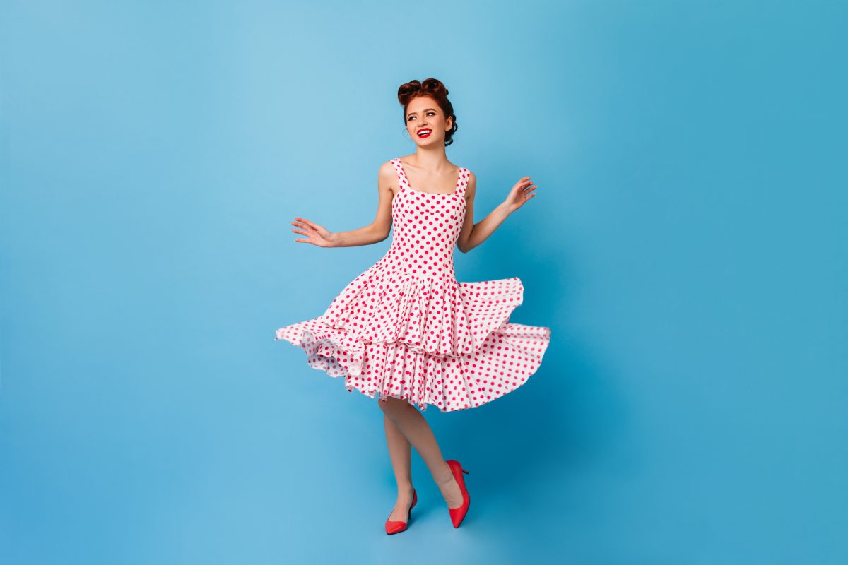 Carefree pinup girl dancing and laughing. Cute young woman in polka-dot dress having fun on blue background.