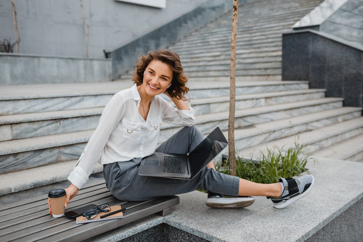 attractive stylish smiling woman working typing on laptop, freelancer sitting in park on stairs drinking coffee, urban city street style, business center, online education