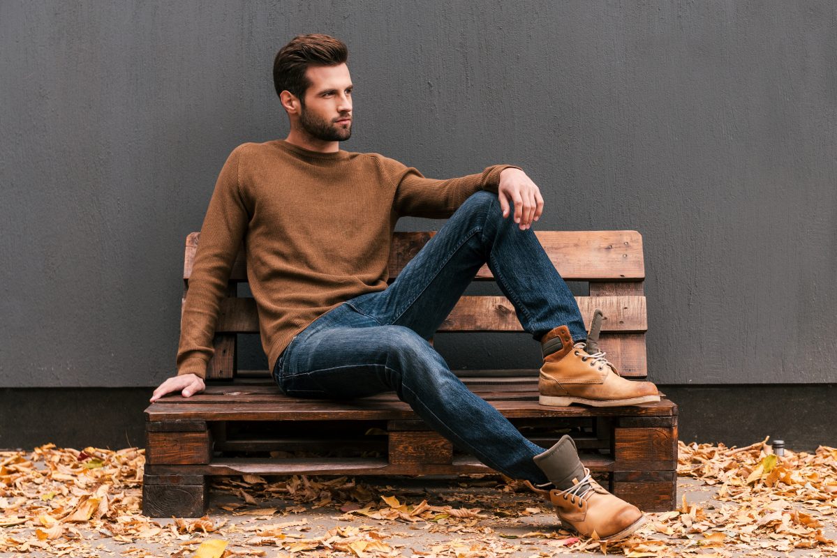 Casually handsome. Handsome young man sitting on the wooden pallet and looking away with grey wall in the background and orange fallen leaves on the floor